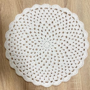Pode incluir: Uma toalha de mesa redonda branca de crochê com uma borda ondulada. A toalha de mesa apresenta um design aberto e rendado com um padrão circular central que irradia para fora. A peça feita à mão é colocada em uma superfície de madeira marrom clara.