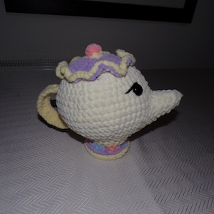 May include: A crocheted teapot in a whimsical design. The teapot is mainly white with a yellow handle and accents of purple, pink, and pastel colours. It has a round body, a spout, and a lid with a pink pom-pom.