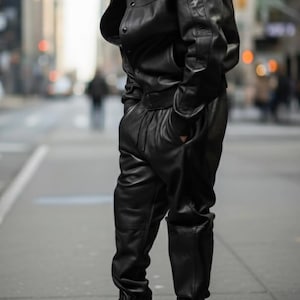 Pu&ograve; includere: Una persona indossa una giacca di pelle nera, pantaloni abbinati e un berretto da baseball nero. L'outfit &egrave; abbinato a scarpe da ginnastica bianche. La persona &egrave; in piedi su una strada cittadina.
