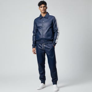 Può includere: Un completo giacca e pantaloni in pelle blu navy. La giacca ha una chiusura con bottoni, un colletto e strisce bianche sulle maniche. I pantaloni hanno una vita con coulisse e strisce bianche sui lati. Le scarpe da ginnastica bianche completano l'outfit.