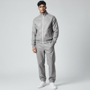 Può includere: Una tuta grigio chiaro con giacca con zip e pantaloni abbinati. La giacca ha un collo alto e finiture bianche lungo le cuciture. I pantaloni hanno un design a gamba dritta con finiture bianche. L'outfit è abbinato a scarpe da ginnastica bianche.