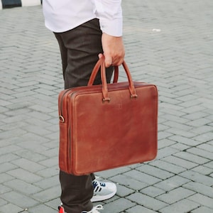 May include: A brown leather briefcase with a top handle and a zipper closure. The briefcase is being held by a person wearing a white shirt and brown pants.