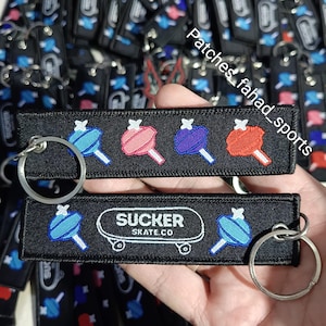 May include: Black embroidered keychains featuring colorful lollipop designs. One keychain has the text "SUCKER SKATE.CO" with a skateboard graphic. The keychains have silver keyrings and are likely for skate enthusiasts.