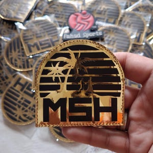 May include: Gold and black patch with a tropical sunset design. The patch features palm trees, silhouettes of people, and the letters "MSH". The patch is likely for sports or team apparel.