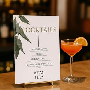 Op de afbeelding: Een cocktailmenu met het woord "COCKTAILS" bovenaan, met dranknamen en ingrediënten. Een feloranje cocktail met een sinaasappelschijf als garnering staat naast het menu op een bar.