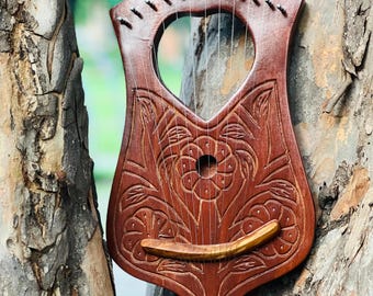 May include: A handcrafted, wooden lyre with a carved floral design. The instrument has a heart-shaped sound hole and a curved bridge. The lyre is a reddish-brown color and has strings stretched across the body.
