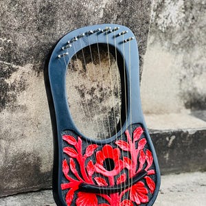May include: A dark blue lyre with a red floral design carved into the soundboard. The instrument has multiple strings and tuning pegs. The lyre is resting against a textured gray surface.