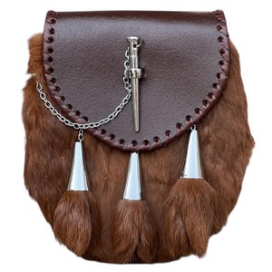 May include: A brown leather sporran with a fur body and silver accents. The sporran features a flap with decorative stitching, a silver chain, and three silver tassels with fur. A silver pin is attached to the flap.