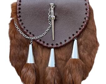 May include: A brown leather sporran with a fur body and silver accents. The sporran features a flap with decorative stitching, a silver chain, and three silver tassels with fur. A silver pin is attached to the flap.