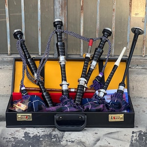 Puede incluir: Una maleta de transporte de cuero negro con un cierre plateado y un asa. Dentro de la maleta hay cuatro gaitas negras con detalles plateados y un chanter blanco y negro.