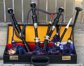 May include: A black leather carrying case with a silver latch and a handle. Inside the case are four black bagpipes with silver accents and a white and black chanter.