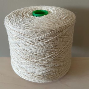 Puede incluir: Una madeja de hilo de color natural enrollada en un carrete de cartón con un centro de plástico verde.