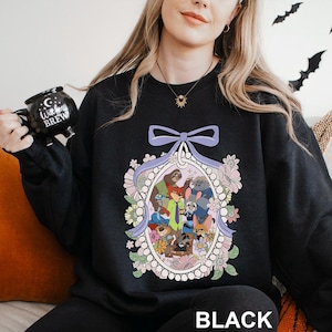 May include: Black sweatshirt with a colourful oval design featuring cartoon animal characters, surrounded by flowers and a light purple bow. The word "BLACK" is printed at the bottom. A black mug with the text "Witches Brew" is held in the image.