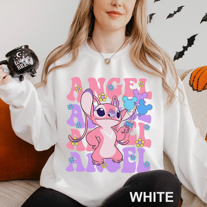 Peut inclure: Sweat-shirt blanc avec un personnage de dessin anim&eacute; rose et le mot "ANGEL" dans une police r&eacute;tro. Le motif comprend des fleurs et des accents bleus et violets. Le mot "WHITE" est imprim&eacute; en bas.
