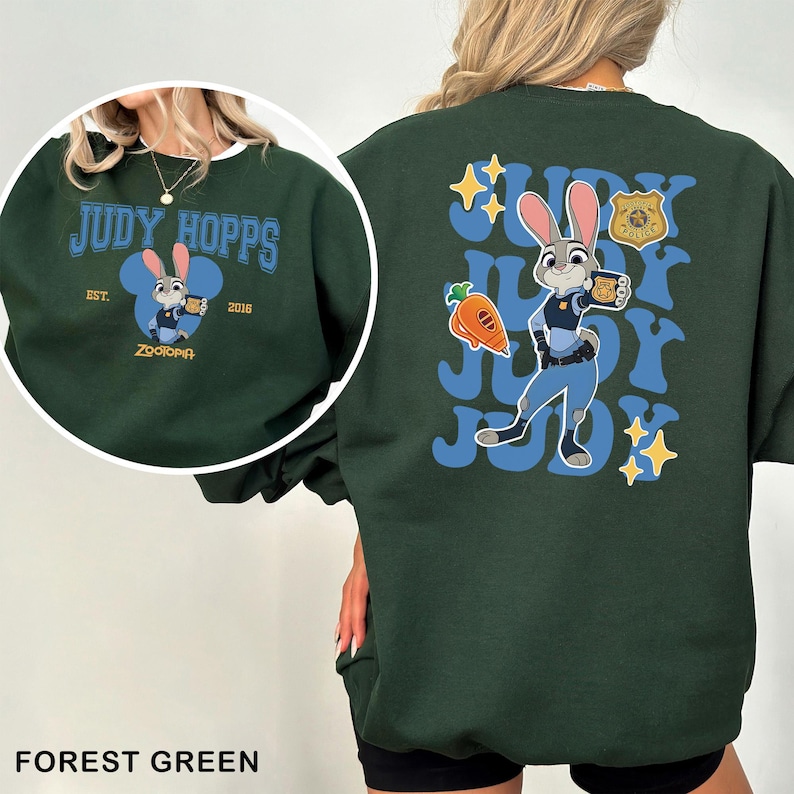 Puede incluir: Sudadera verde bosque con una imagen de dibujos animados de Judy Hopps de Zootopia. La parte delantera tiene el texto "JUDY HOPPS" y "EST. 2016 Zootopia". La parte trasera muestra una imagen m&aacute;s grande de Judy Hopps con su uniforme de polic&iacute;a, con el texto "JUDY" repetido.