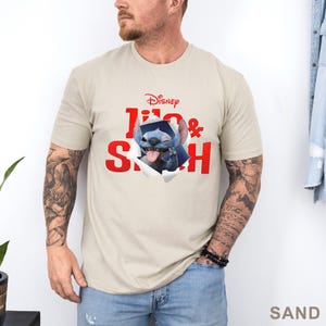 May include: Sand-colored t-shirt featuring the Disney logo and the words "Lilo & Stitch" in red. The graphic shows Stitch bursting through the shirt. The shirt is made of a soft material.