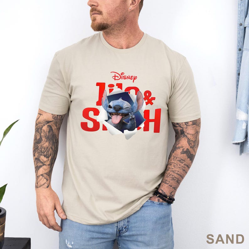 May include: Sand-colored t-shirt featuring the Disney logo and the words "Lilo & Stitch" in red. The graphic shows Stitch bursting through the shirt. The shirt is made of a soft material.