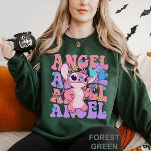 Peut inclure: Sweat-shirt vert for&ecirc;t avec le mot "ANGEL" dans une police r&eacute;tro et un personnage de dessin anim&eacute; rose. Le personnage tient une fleur bleue. Le sweat-shirt porte &eacute;galement l'inscription "FOREST GREEN" en bas.
