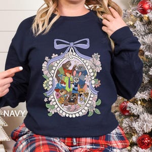 May include: Navy blue sweatshirt with a framed cartoon illustration of animal characters. The oval frame is decorated with flowers and a light purple bow. The word "NAVY" is printed on the left side of the sweatshirt.