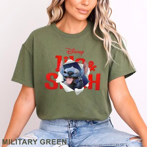 May include: Military green t-shirt featuring the Disney characters Lilo & Stitch. The design shows Stitch bursting through a torn paper effect, with the Disney logo above. The text "Lilo & Stitch" is in red.