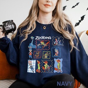 Puede incluir: Sudadera azul marino con la palabra "Zootopia" e ilustraciones de personajes de dibujos animados en marcos coloridos. La sudadera tiene cuello redondo y mangas largas. La palabra "NAVY" est&aacute; impresa en la esquina inferior derecha.