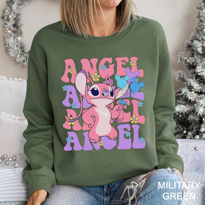 Peut inclure: Sweat-shirt vert militaire avec un personnage de dessin anim&eacute; rose tenant des fleurs, avec le mot "ANGEL" en lettres roses et violettes. Le motif comprend des fleurs bleues et jaunes.