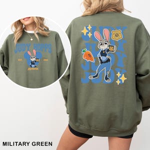Puede incluir: Sudadera verde militar con un dise&ntilde;o de Judy Hopps. La parte delantera muestra "Judy Hopps" en azul, una ilustraci&oacute;n de conejo y el texto "Zootopia". La parte trasera presenta a Judy Hopps con uniforme de polic&iacute;a, una zanahoria y estrellas.