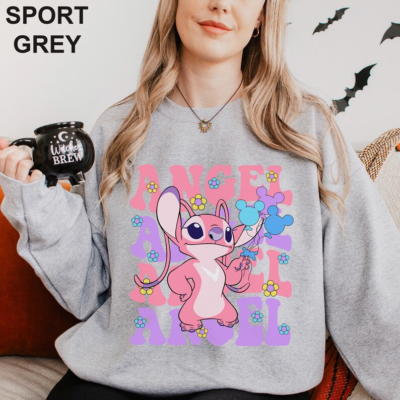 Peut inclure: Sweat-shirt gris sport avec un personnage de dessin anim&eacute; rose tenant des ballons bleus. Le mot "ANGEL" est imprim&eacute; en rose et violet, entour&eacute; de fleurs. Le sweat-shirt est de style col rond.