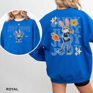 Puede incluir: Sudadera azul real con una imagen de dibujos animados de Judy Hopps de Zootopia. La parte delantera tiene el texto "JUDY HOPPS" con una imagen de dibujos animados de Judy. La parte trasera tiene una imagen m&aacute;s grande de Judy con el texto "JUDY" repetido.