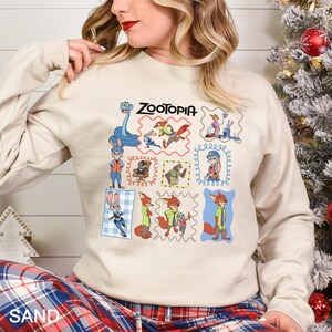 Puede incluir: Una sudadera de color arena con un gr&aacute;fico colorido de personajes de la pel&iacute;cula animada "Zootopia". El dise&ntilde;o incluye varios personajes en ilustraciones enmarcadas. La palabra "Zootopia" est&aacute; impresa encima de las ilustraciones.