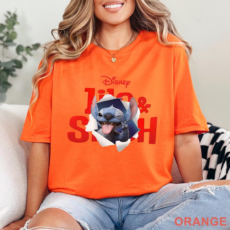 May include: Orange t-shirt featuring the Disney logo and the words "Lilo & Stitch" in red. A graphic of Stitch, the blue alien, is breaking through the shirt. The word "ORANGE" is printed at the bottom of the shirt.