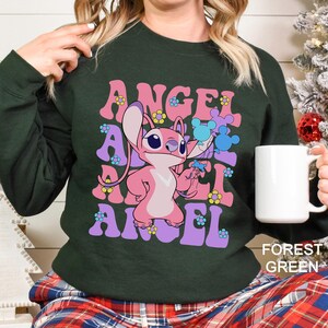 Peut inclure: Sweat-shirt vert for&ecirc;t avec le mot "ANGEL" en rose et violet dans une police r&eacute;tro, et un personnage de dessin anim&eacute; rose tenant des objets bleus. Le sweat-shirt a un col rond et des manches longues. Le texte "FOREST GREEN" est en bas.
