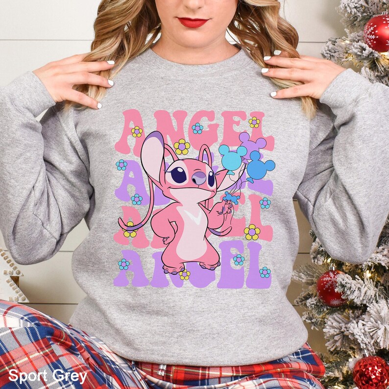 Peut inclure: Sweat-shirt gris sport avec un personnage de dessin anim&eacute; rose tenant des ballons bleus. Le mot "ANGEL" est r&eacute;p&eacute;t&eacute; en rose et violet, avec des fleurs jaunes. Le sweat-shirt est de style d&eacute;contract&eacute;.