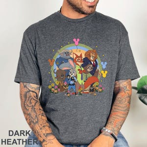 Puede incluir: Camiseta gris jaspeado oscuro con una colorida ilustraci&oacute;n de dibujos animados de los personajes de Zootopia, incluyendo a Judy Hopps y Nick Wilde. El dise&ntilde;o presenta flores, iconos de Mickey Mouse y el texto "DARK HEATHER" en la parte inferior.