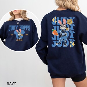Puede incluir: Sudadera azul marino con Judy Hopps de Zootopia. El dise&ntilde;o frontal incluye el texto "JUDY HOPPS" sobre una imagen de dibujos animados de Judy. El dise&ntilde;o trasero muestra a Judy con su uniforme de polic&iacute;a, con la palabra "JUDY" repetida.