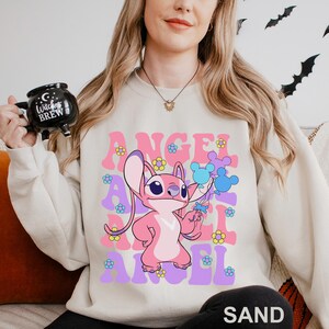 Peut inclure: Sweat-shirt couleur sable avec un personnage de dessin anim&eacute; rose tenant des ballons bleus. Le mot "ANGEL" est imprim&eacute; en rose et violet, avec des motifs de fleurs. Le sweat-shirt est de style d&eacute;contract&eacute;.