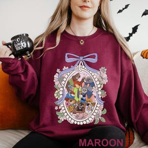 May include: Maroon sweatshirt with a floral oval frame containing cartoon characters. The frame is surrounded by flowers and a light purple ribbon. The word "MAROON" is printed at the bottom of the sweatshirt.