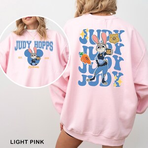 Puede incluir: Sudadera rosa claro con una imagen de dibujos animados de Judy Hopps de Zootopia. La parte delantera tiene un dise&ntilde;o circular con el texto "JUDY HOPPS" y "Zootopia". La parte trasera tiene un dise&ntilde;o m&aacute;s grande con Judy Hopps y la palabra "JUDY".