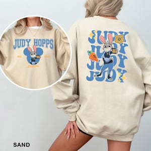 Puede incluir: Sudadera color arena con una imagen de dibujos animados de Judy Hopps de Zootopia. El dise&ntilde;o frontal incluye el texto "Judy Hopps" y una imagen de dibujos animados de Judy. El dise&ntilde;o trasero presenta a Judy Hopps con uniforme de polic&iacute;a con el texto "Judy".