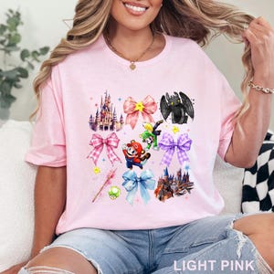 May include: Light pink t-shirt featuring a colorful graphic design. The design includes illustrations of castles, bows, and cartoon characters. The text "LIGHT PINK" is printed at the bottom of the shirt.