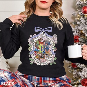 May include: Black crew neck sweatshirt with a floral oval design featuring cartoon characters. The design includes a light purple bow at the top and is surrounded by flowers in pink, white, and green. The word "BLACK" is printed at the bottom.