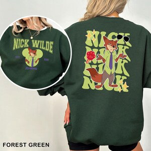 Puede incluir: Sudadera verde bosque con un gr&aacute;fico de Nick Wilde de Zootopia. La parte delantera tiene un dise&ntilde;o circular con texto y una ilustraci&oacute;n de zorro. La parte trasera muestra un gr&aacute;fico m&aacute;s grande de Nick Wilde con gafas de sol y texto. La sudadera es de cuello redondo.