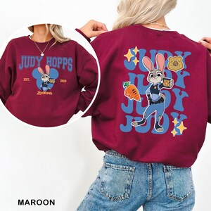 Puede incluir: Sudadera granate con Judy Hopps de Zootopia. El dise&ntilde;o frontal incluye el texto "JUDY HOPPS" y una imagen de dibujos animados de Judy. La parte trasera muestra un gr&aacute;fico m&aacute;s grande de Judy Hopps con el texto "JUDY".