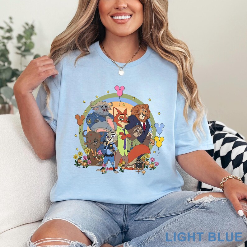 Puede incluir: Camiseta azul claro con una ilustraci&oacute;n de dibujos animados de personajes de animales. El dise&ntilde;o incluye un arco&iacute;ris, flores y el texto "LIGHT BLUE" en la parte inferior. La camiseta tiene mangas cortas y cuello redondo.