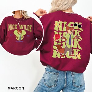 Puede incluir: Sudadera granate con un dise&ntilde;o de personaje de zorro de dibujos animados. La parte delantera de la sudadera tiene el texto "Nick Wilde" y un gr&aacute;fico de zorro de dibujos animados. La parte trasera presenta un zorro de dibujos animados m&aacute;s grande con gafas de sol.