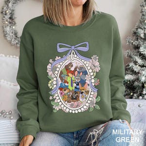 May include: Military green sweatshirt with a cartoon illustration of animal characters in an oval frame, surrounded by flowers and a purple bow. The sweatshirt is a solid military green colour.