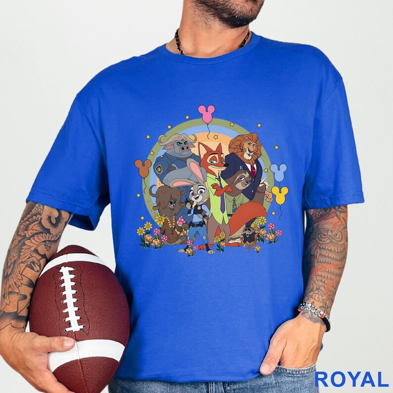 Puede incluir: Camiseta azul real con un gr&aacute;fico de dibujos animados de personajes de animales, incluyendo un zorro, un le&oacute;n y un conejo. El gr&aacute;fico est&aacute; en un dise&ntilde;o circular con flores y globos. La camiseta la sostiene una persona con un bal&oacute;n de f&uacute;tbol americano.