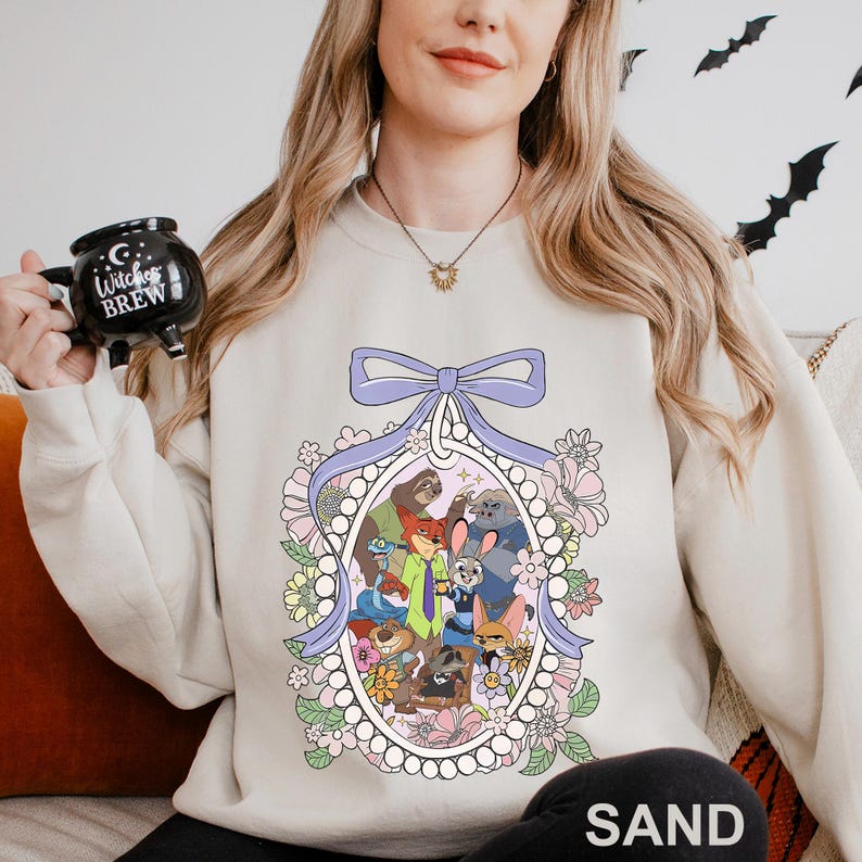May include: Sand-coloured sweatshirt with a cartoon illustration of animal characters in an oval frame, surrounded by flowers and a light purple bow. The word "SAND" is printed at the bottom.