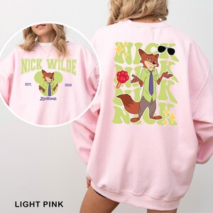 Puede incluir: Sudadera rosa claro con una ilustraci&oacute;n de dibujos animados de Nick Wilde de Zootopia. La parte delantera muestra "Nick Wilde" y "Zootopia". La parte trasera presenta una imagen m&aacute;s grande de Nick Wilde con gafas de sol y un helado, y el texto "Nick Wilde".