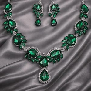 May include: A dazzling emerald green jewelry set, including a necklace and matching earrings. The necklace features large, teardrop-shaped stones and smaller accent stones, all set in a silver-toned metal. The earrings mirror the necklace design. The set is displayed on a gray satin surface.
